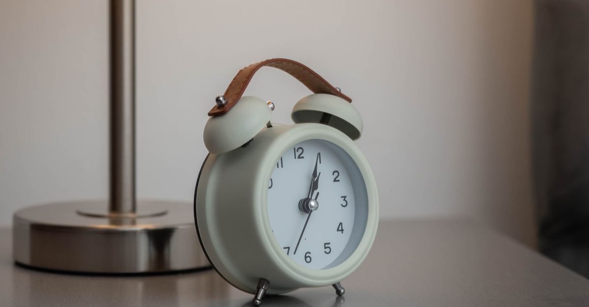 A classic analogue alarm clock beside a glowing lamp on a bedside table.