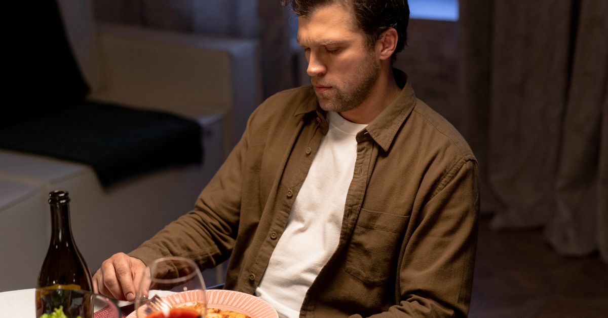 Man sitting alone at a dinner table with a meal and a glass of wine, looking thoughtful